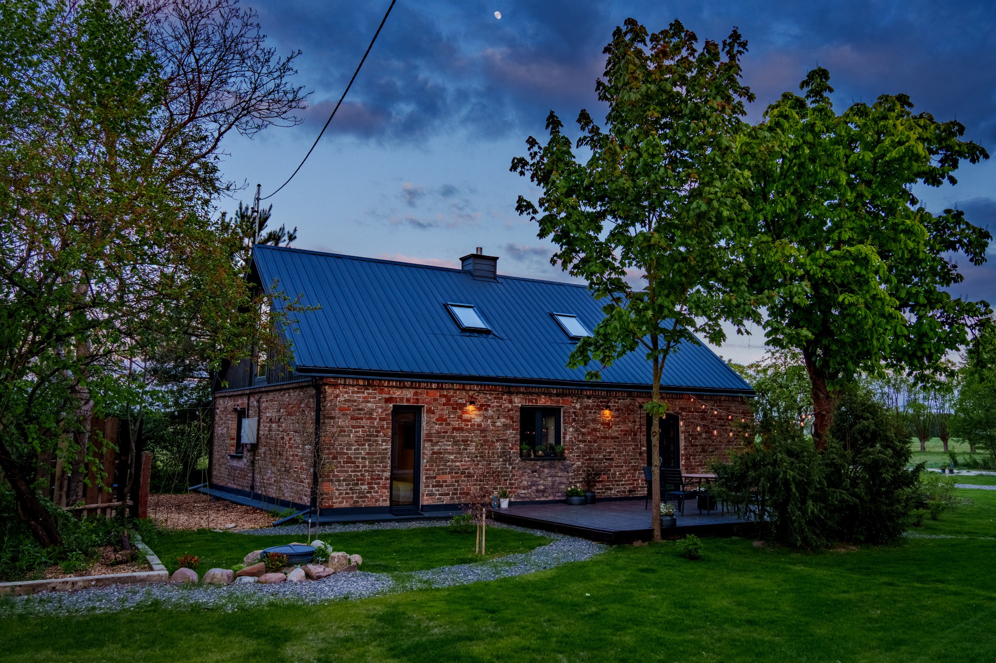 Ceglany dom z ciemnym dachem i oknami połaciowymi, otoczony zielenią, sfotografowany o zmierzchu w Warszawie. Widoczny ogród i elementy architektury.