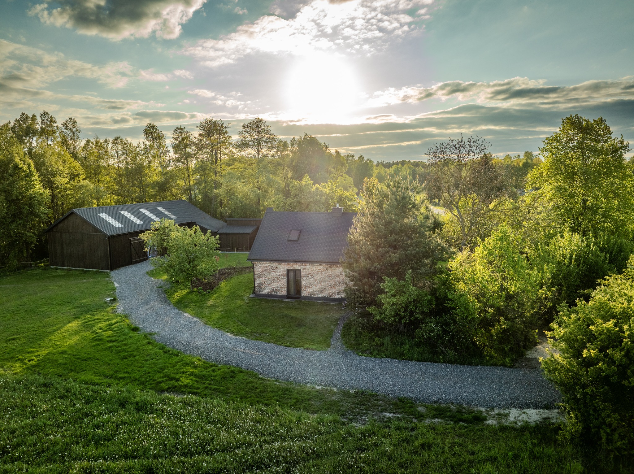Panoramiczny widok z lotu ptaka na kamienny dom z ciemnym dachem i budynki gospodarcze, otoczony zielenią i krętą ścieżką żwirową w słoneczny dzień.