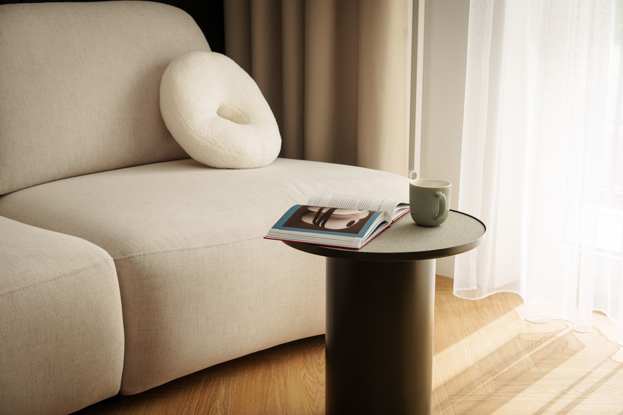 Jasny, minimalistyczny salon: beżowa sofa z okrągłą poduszką, stolik z książką i kubkiem, parkiet w jodełkę, delikatna firanka wpadająca w jasne światło.