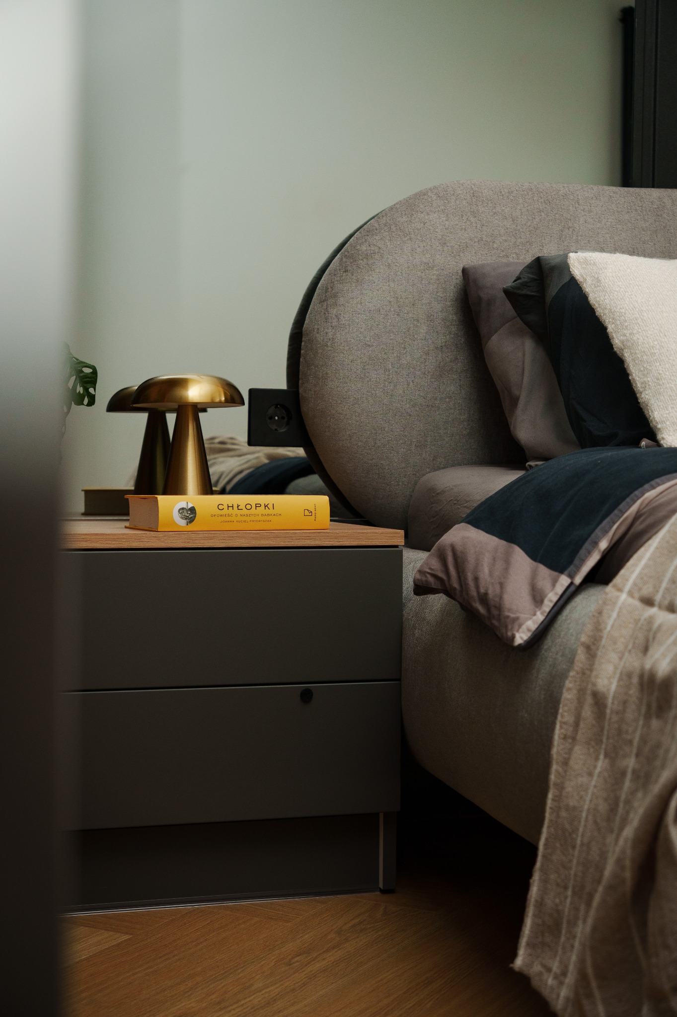 Sypialnia w stonowanych barwach: szare łóżko z okrągłym zagłówkiem, drewniana szafka nocna z książką 'Chłopki' i złotą lampką. Przytulne i eleganckie wnętrze.