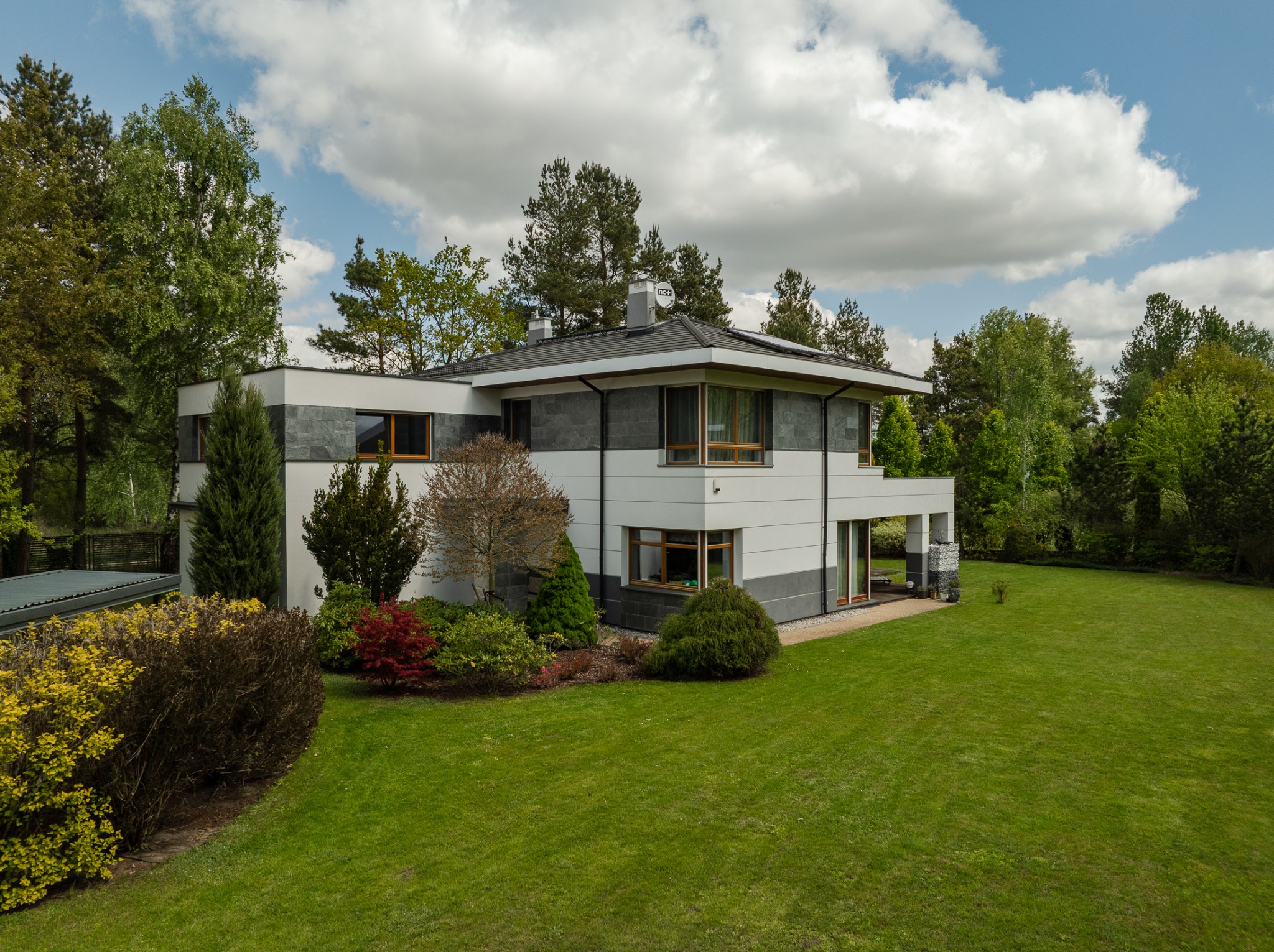 Dwukondygnacyjny dom z szarą elewacją i dużymi oknami, otoczony zielonym trawnikiem i drzewami, sfotografowany w słoneczny dzień z lekkiej perspektywy.