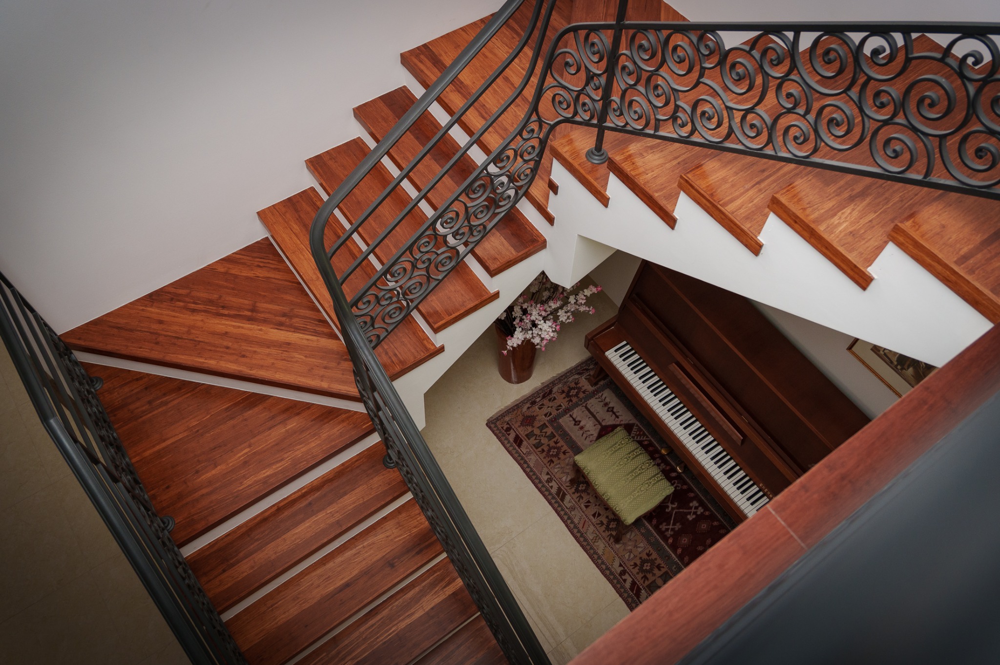 Spiralne schody z drewnianymi stopniami i ozdobną, kutą balustradą. W tle pianino, dywan i wazon z kwiatami. Ujęcie z góry, podkreślające detale wykończenia.