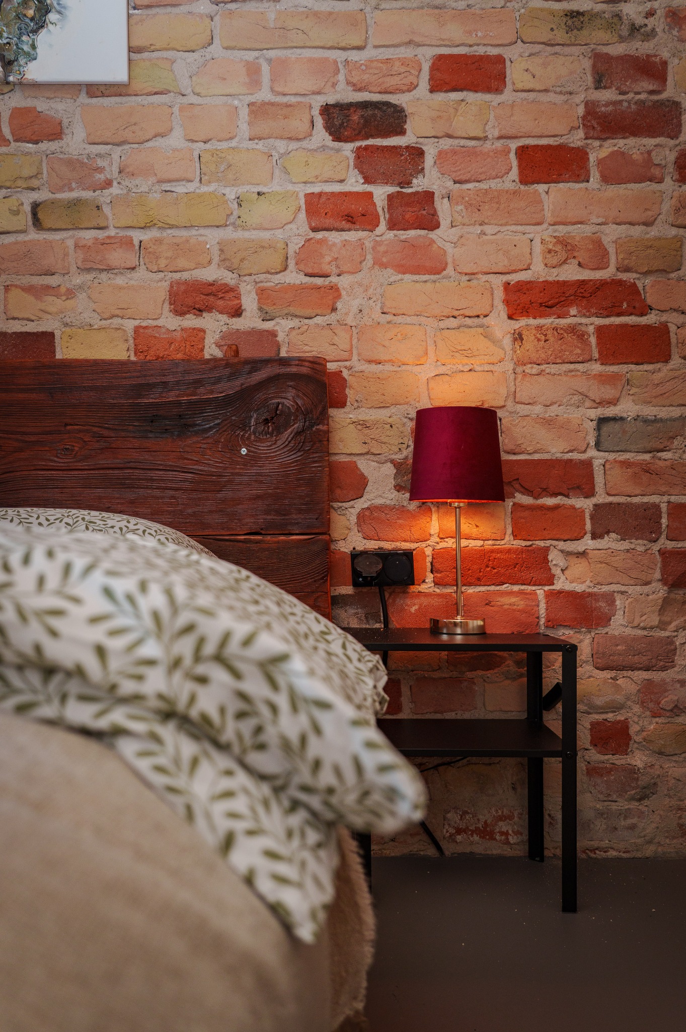 Sypialnia z ceglaną ścianą, drewnianym wezgłowiem łóżka i nocną lampką z bordowym abażurem, oświetlającą fragment pokoju. Widoczne łóżko z pościelą w liście.