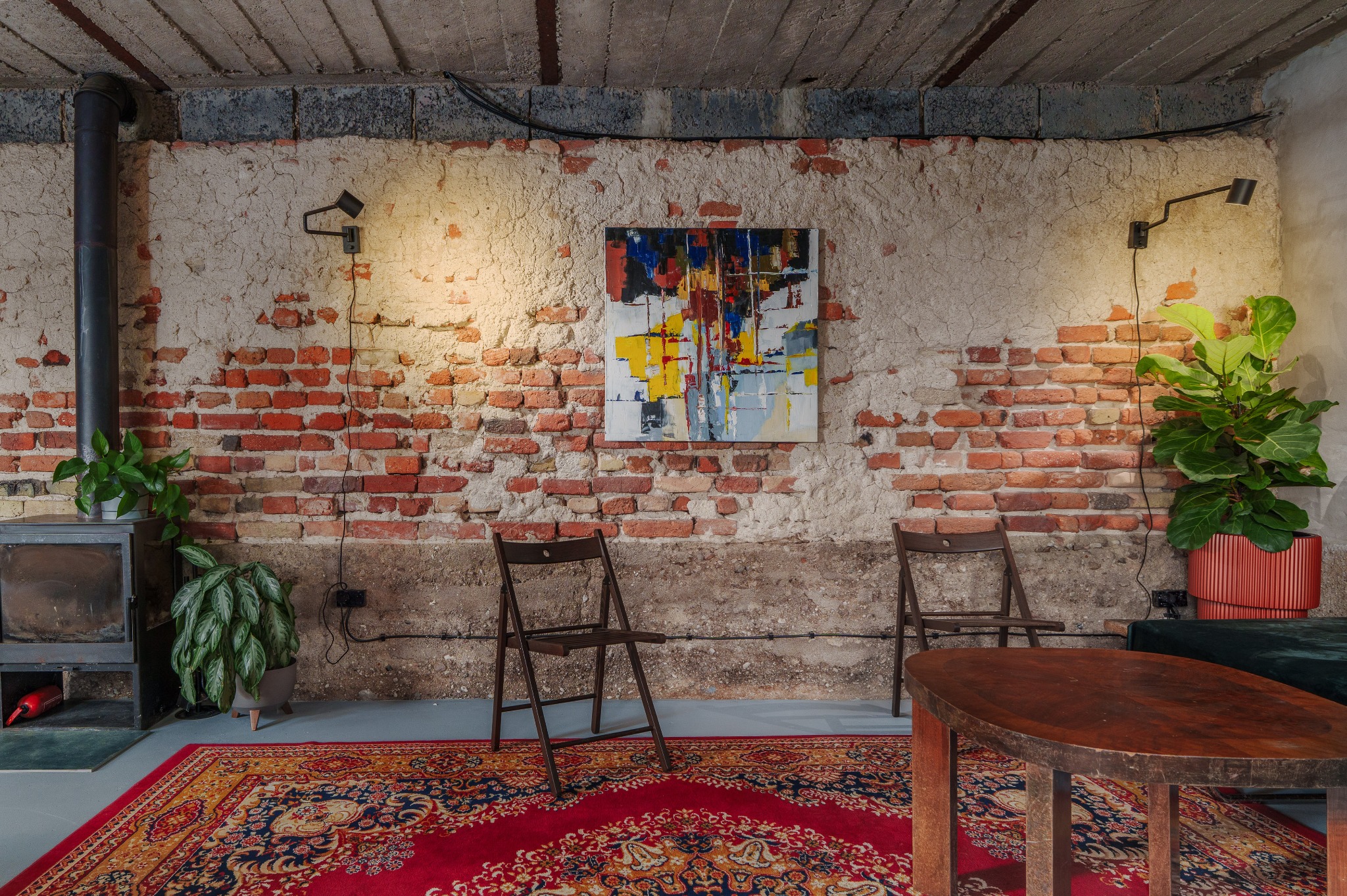 Loftowe wnętrze z ceglaną ścianą, abstrakcyjnym obrazem i drewnianymi krzesłami. Czerwony dywan dodaje ciepła surowej przestrzeni. Oświetlenie punktowe podkreśla detale.