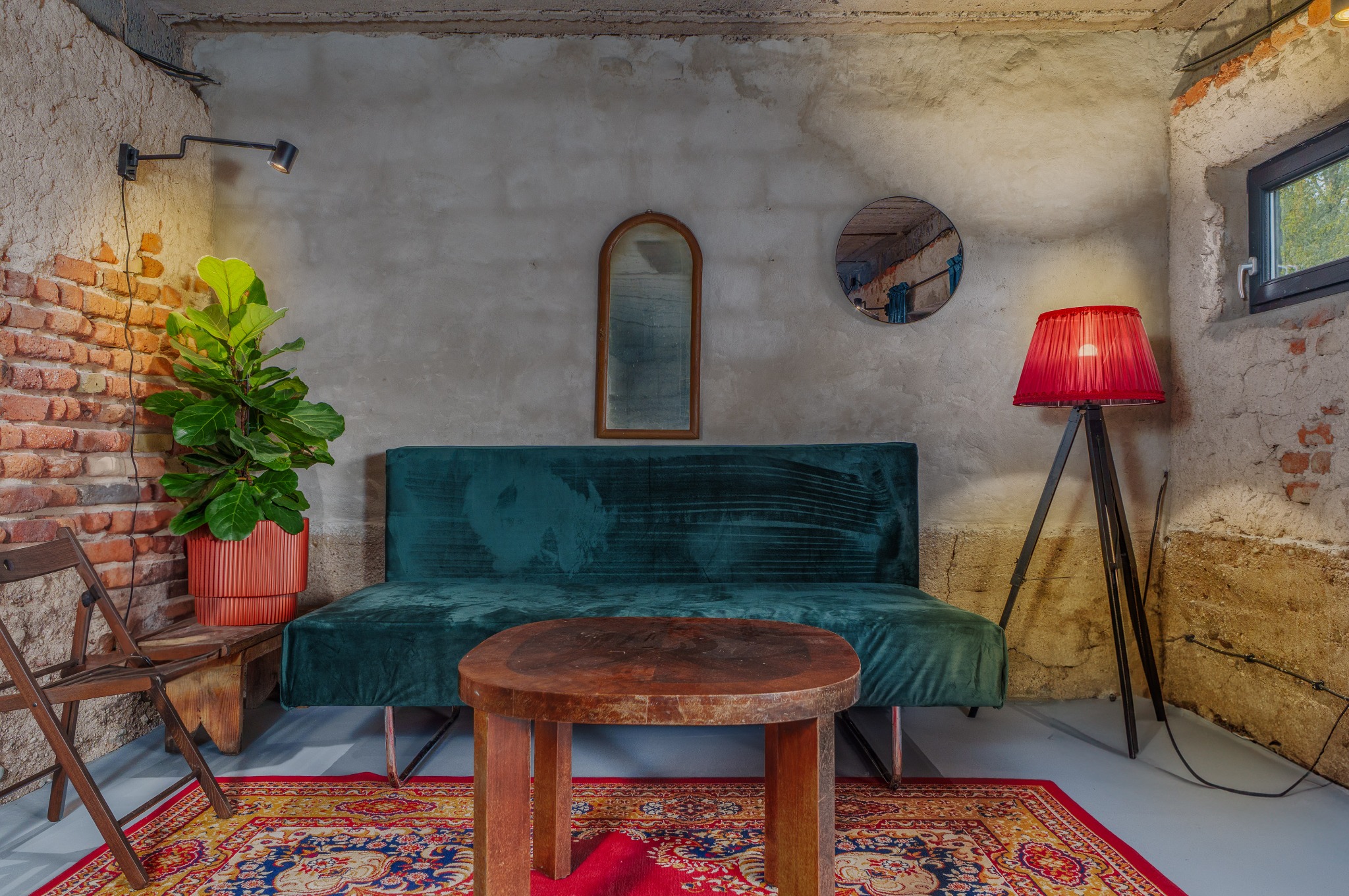 Stylowe wnętrze z zieloną sofą, drewnianym stolikiem i czerwonym dywanem. Na ścianie lustra i lampa. Surowy klimat podkreślają ceglane fragmenty i betonowe ściany.