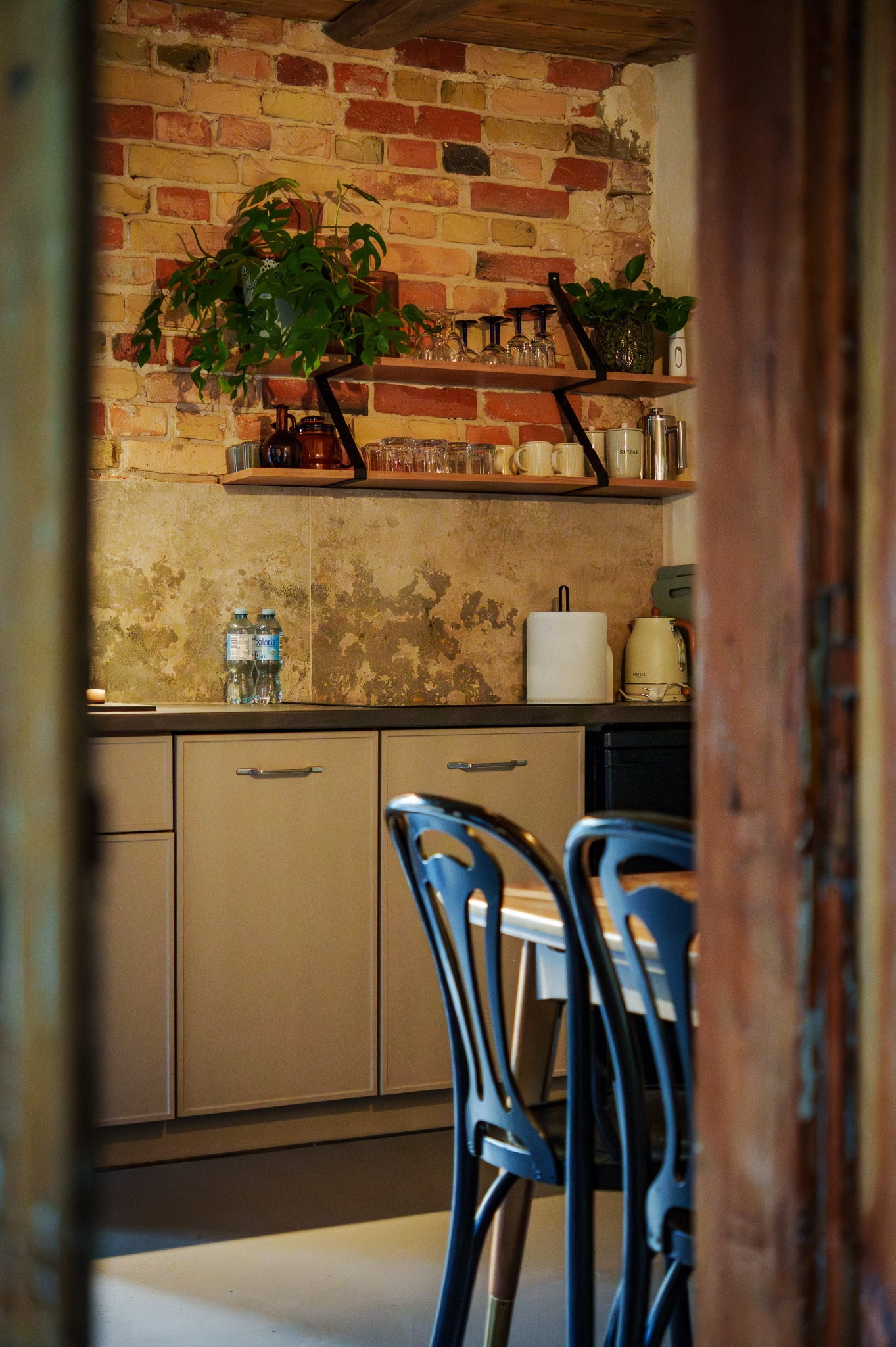 Rustykalna kuchnia z ceglaną ścianą, drewnianymi półkami z roślinami i naczyniami, beżowymi szafkami i czarnymi krzesłami, widziana przez framugę drzwi.