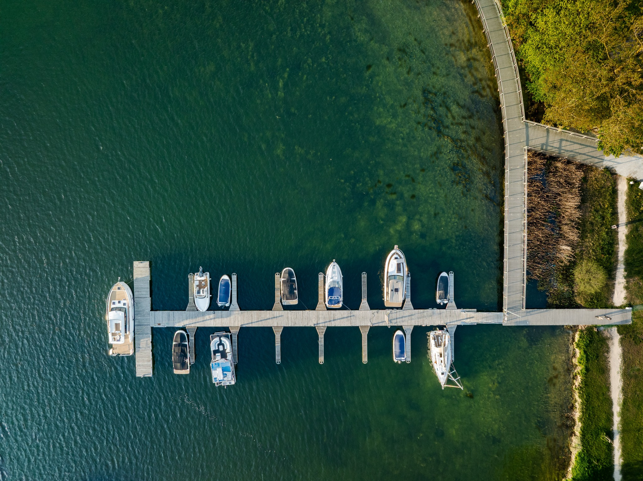 Widok z drona na przystań z drewnianym pomostem, przy którym cumują różnorodne łodzie i jachty, otoczone zieloną wodą i brzegiem z roślinnością.