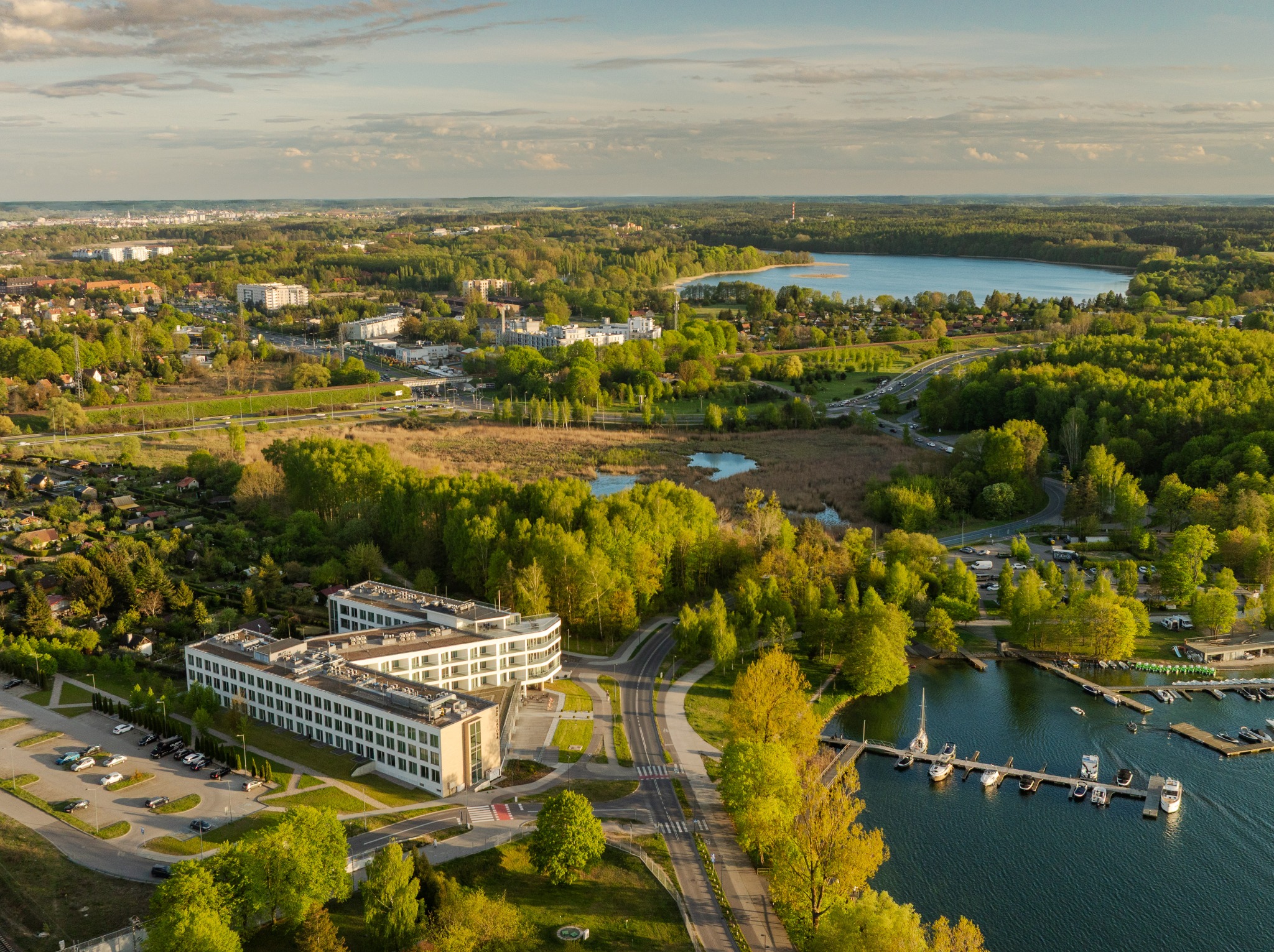Panoramiczny widok z lotu ptaka na nowoczesny budynek biurowy, jezioro z przystanią jachtową i otaczającą zieleń w słoneczny dzień. Ujęcie z drona.