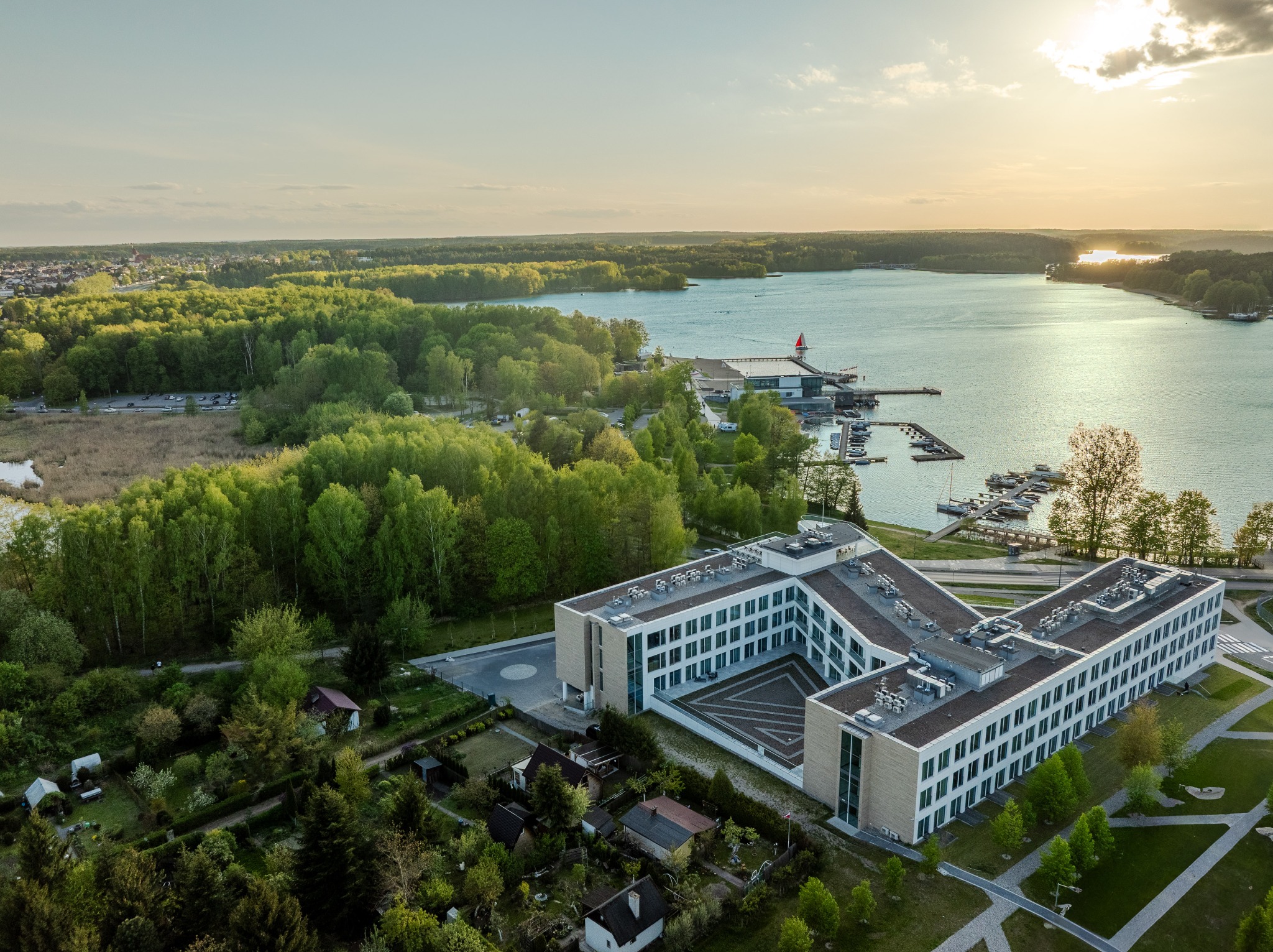 Panorama z lotu ptaka: nowoczesny budynek w kształcie litery V nad jeziorem, otoczony zielenią i małymi domkami, w ciepłym świetle zachodzącego słońca.
