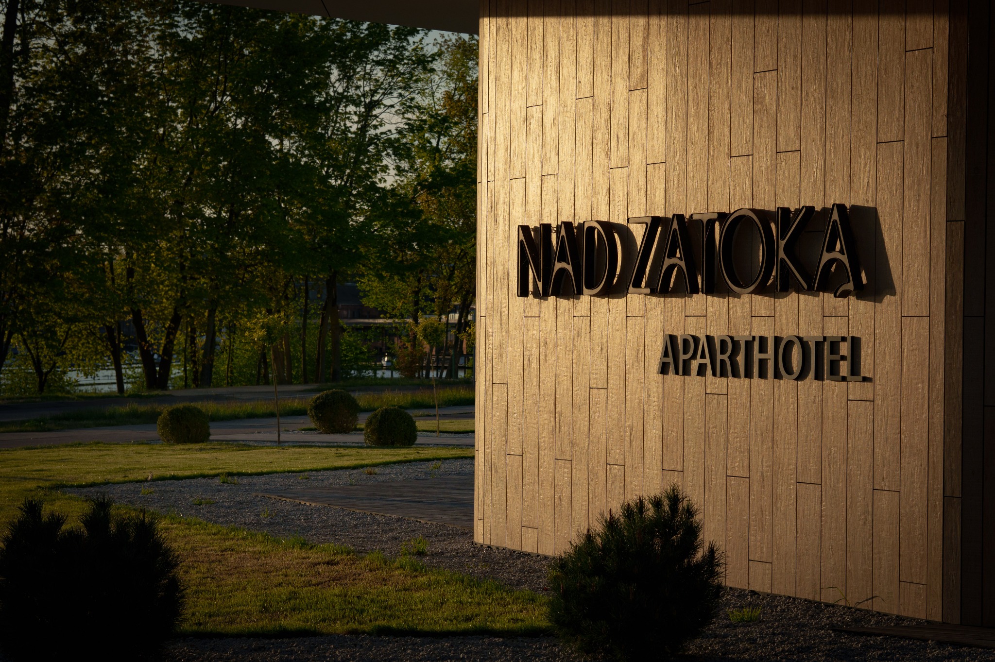 Elewacja budynku z napisem 'NAD ZATOKĄ APARTHOTEL' w ciepłym świetle. W tle zieleń drzew i trawnik z krzewami. Ujęcie z dołu, podkreślające nowoczesny design budynku.