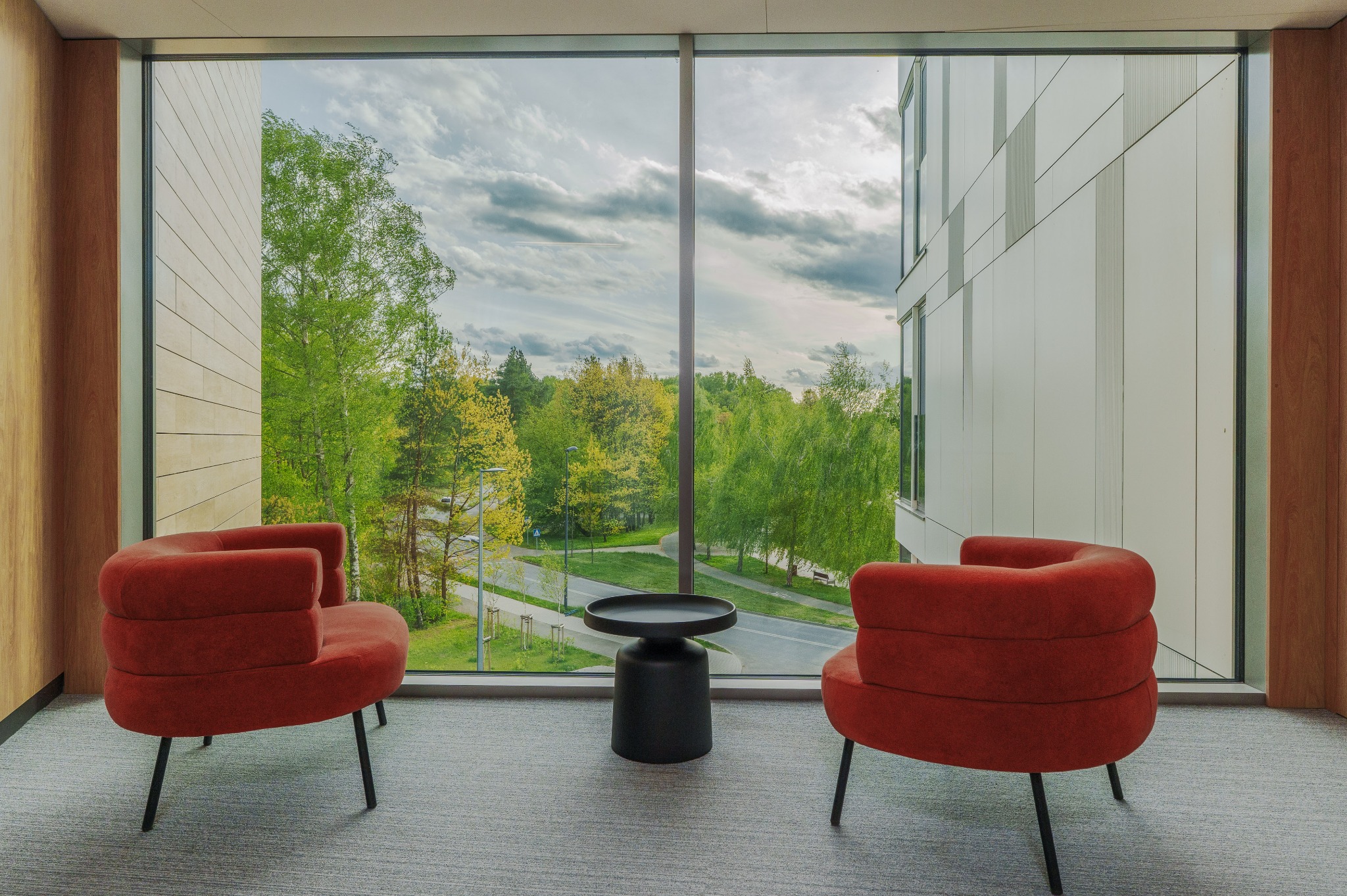 Nowoczesne wnętrze z dwoma fotelami w odcieniach czerwieni i stolikiem kawowym na tle okna z widokiem na zieleń i budynek biurowy.