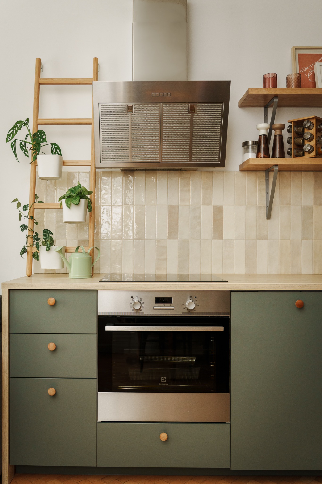 Nowoczesna kuchnia w odcieniach zieleni z drewnianymi akcentami, okapem i półkami. Dekoracyjne rośliny doniczkowe i drabina dodają wnętrzu przytulności. Minimalistyczny design.