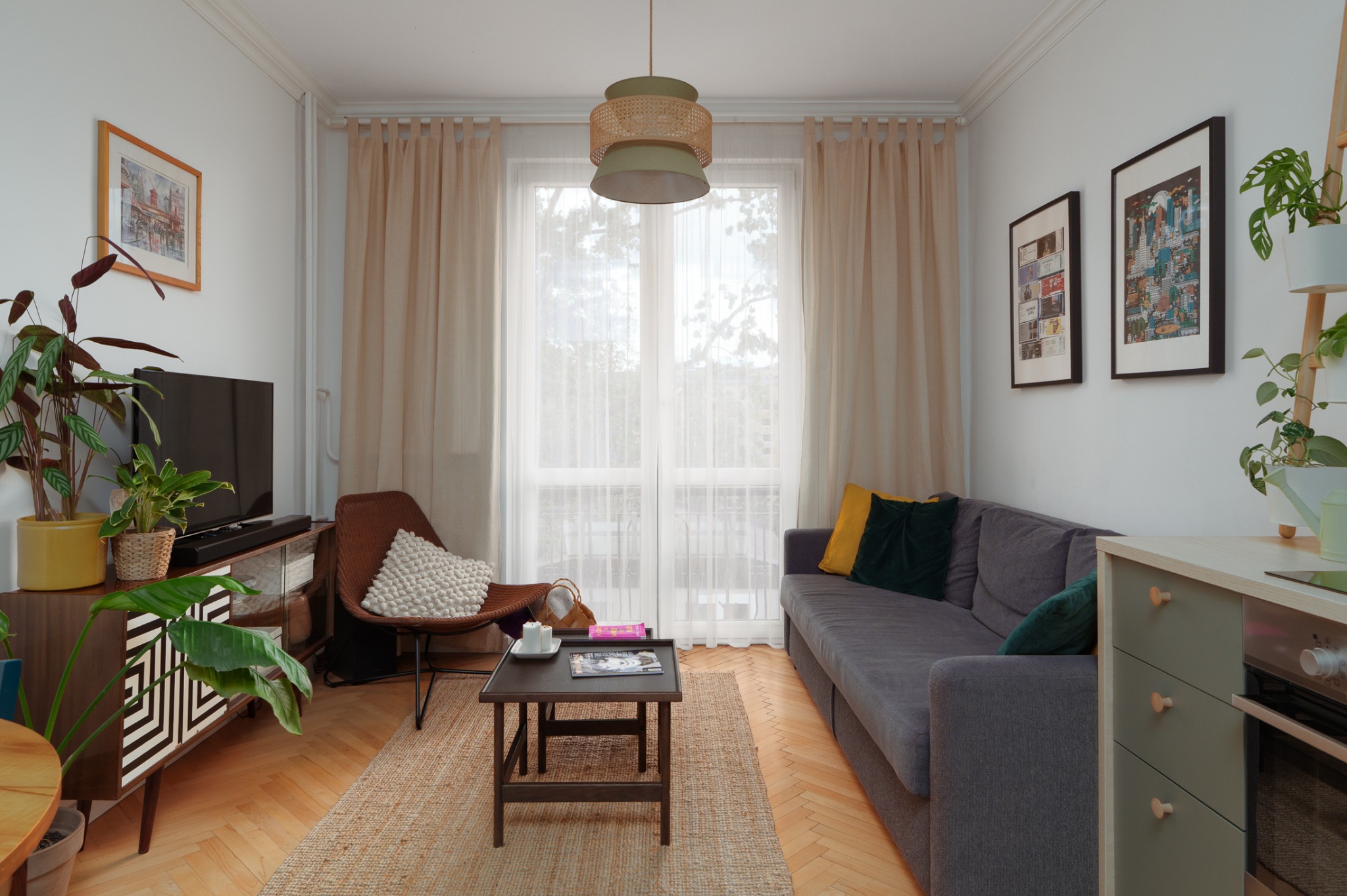 Salon w stylu retro z parkietem w jodełkę, szarą sofą, brązowym fotelem i roślinami doniczkowymi. Na ścianach wiszą grafiki w czarnych ramach.