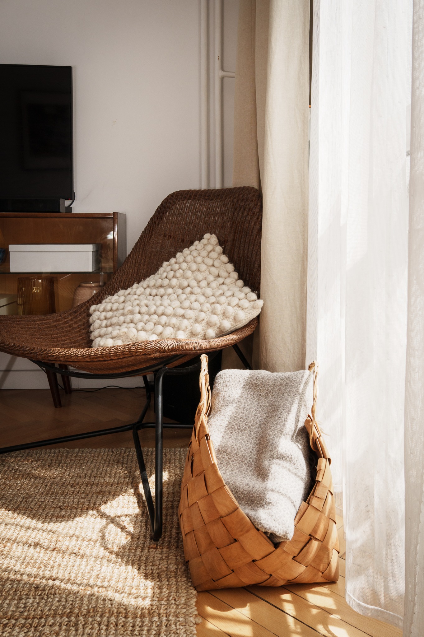 Jasne wnętrze: wiklinowy fotel z poduszką, pleciony kosz z pledem, dywan z juty i zwiewne zasłony w słonecznym świetle. Minimalistyczny wystrój.