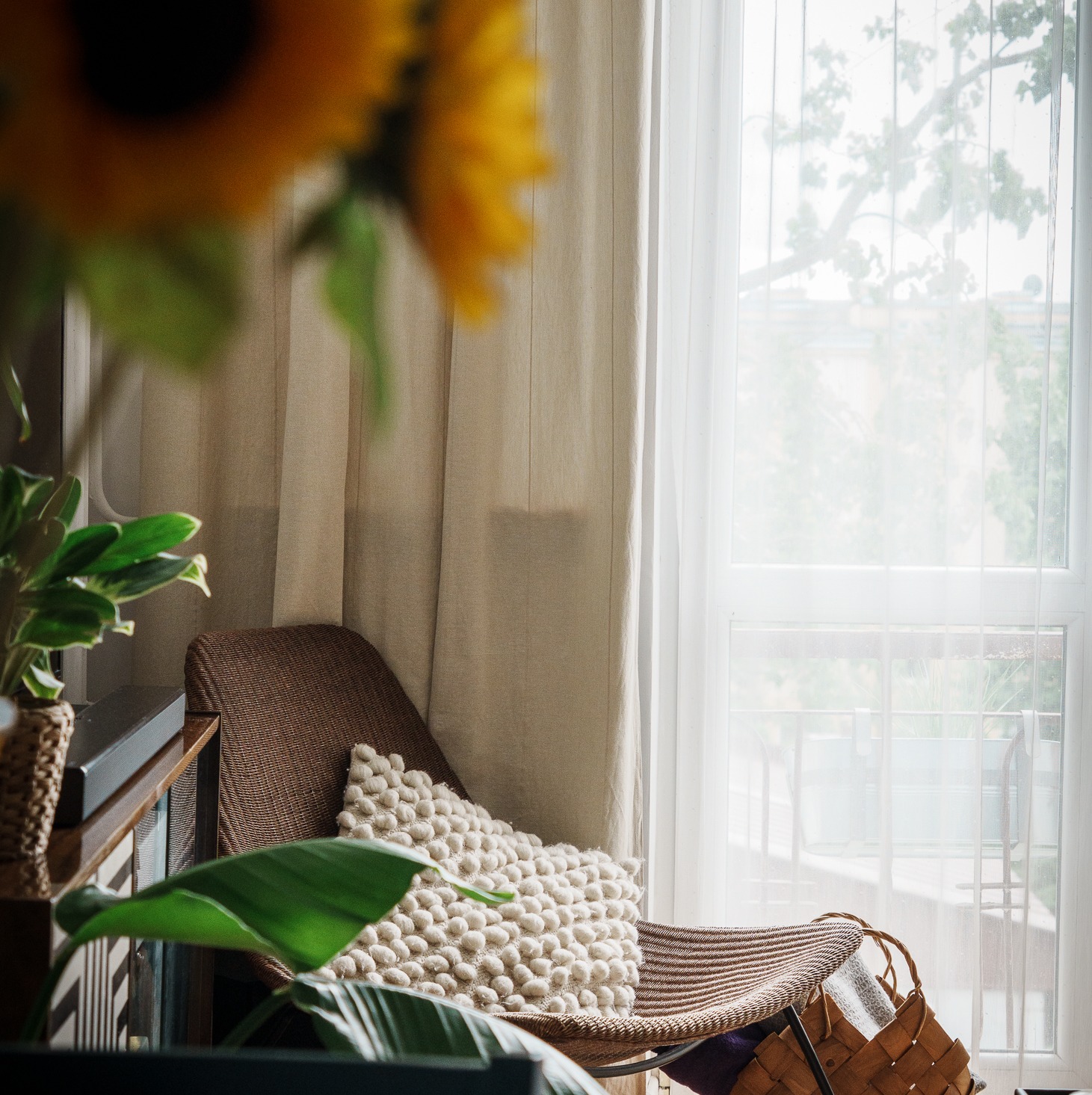 Jasne wnętrze z wiklinowym fotelem, poduszką z pomponami i kwiatami słonecznika. Naturalne światło przez okno z firankami. Spokojna, domowa atmosfera.