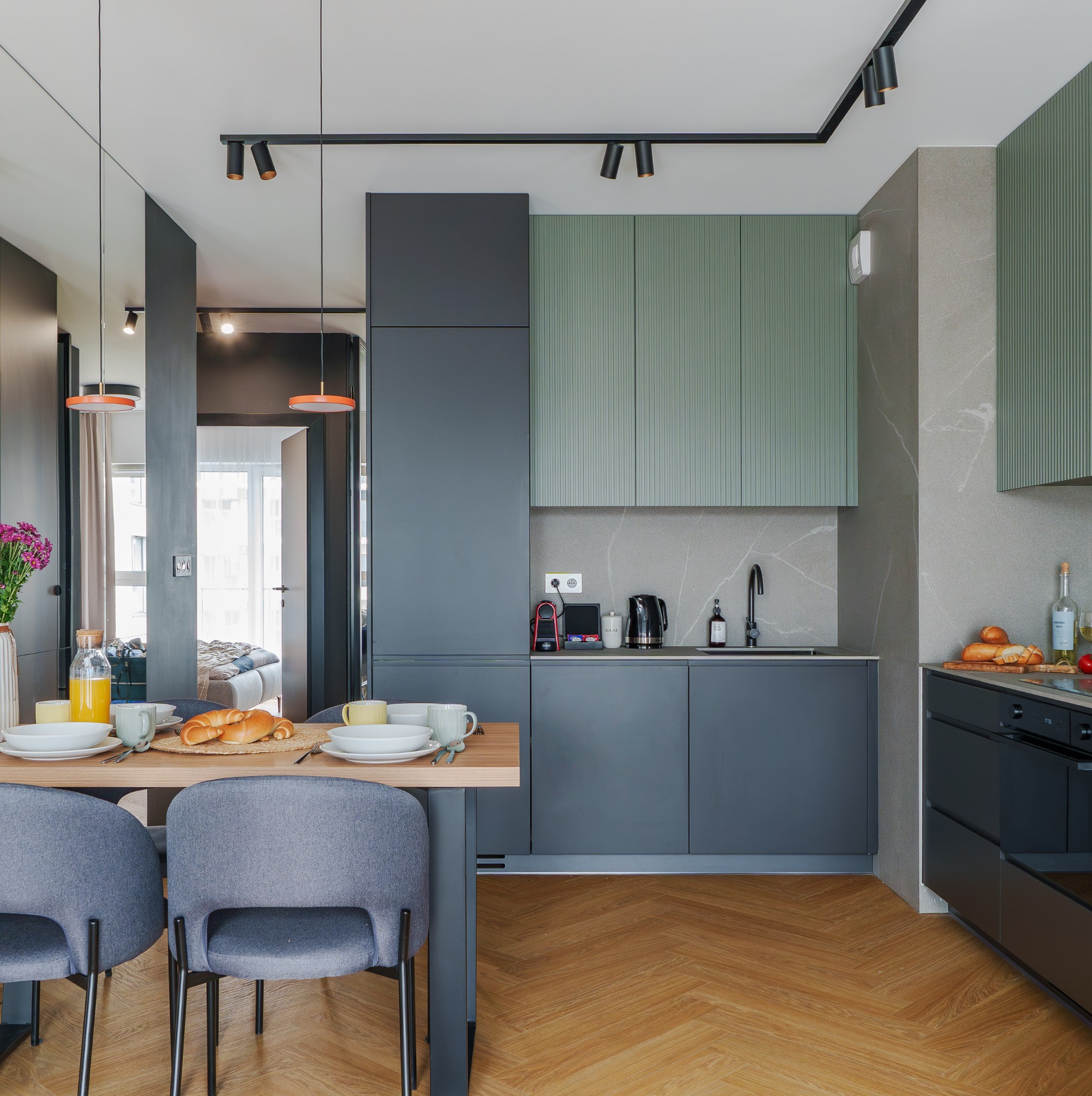 Nowoczesna kuchnia z elementami szarości i zieleni. Stół z zastawą i krzesłami na pierwszym planie, w tle szafki kuchenne i widok na sypialnię.
