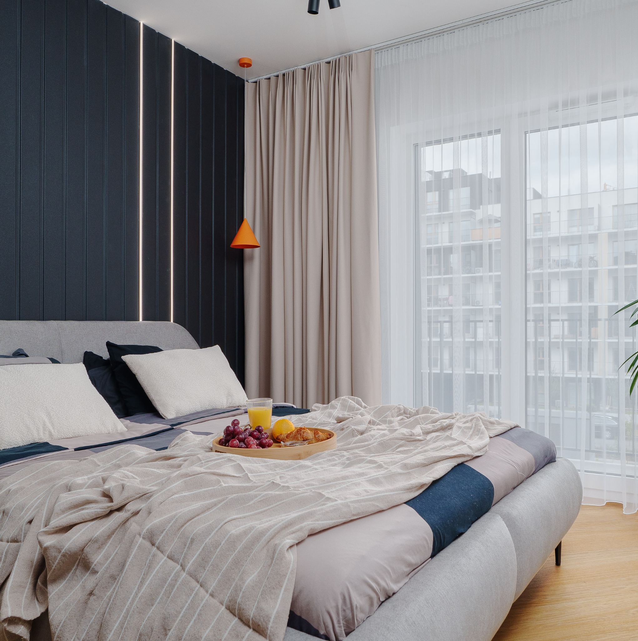 Nowoczesna sypialnia z łóżkiem, śniadaniem na tacy i oknem z widokiem na miasto. Czarna ściana z pionowymi paskami LED. Beżowe zasłony i pomarańczowa lampa.