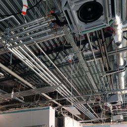 Visma-Tech - Widok na sufit w trakcie instalacji: liczne rury, kanały wentylacyjne, kable i elementy mocujące, z widocznymi fragmentami konstrukcji budynku.