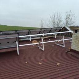 Visma-Tech - Montaż paneli fotowoltaicznych na brązowym, blaszanym dachu domu jednorodzinnego w pochmurny dzień, widoczne elementy konstrukcyjne i fragmenty kartonów po opakowaniach.