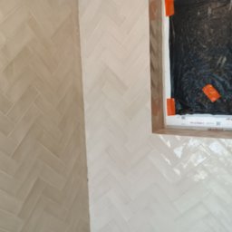 Visma-Tech - Ściana wyłożona błyszczącymi, kremowymi kafelkami w jodełkę, z widocznym oknem zabezpieczonym folią i taśmą malarską podczas prac wykończeniowych.