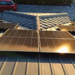 Panele fotowoltaiczne zamontowane na blaszanym dachu z widocznym odbiciem słońca, w tle czerwony samochód i fragment boiska.