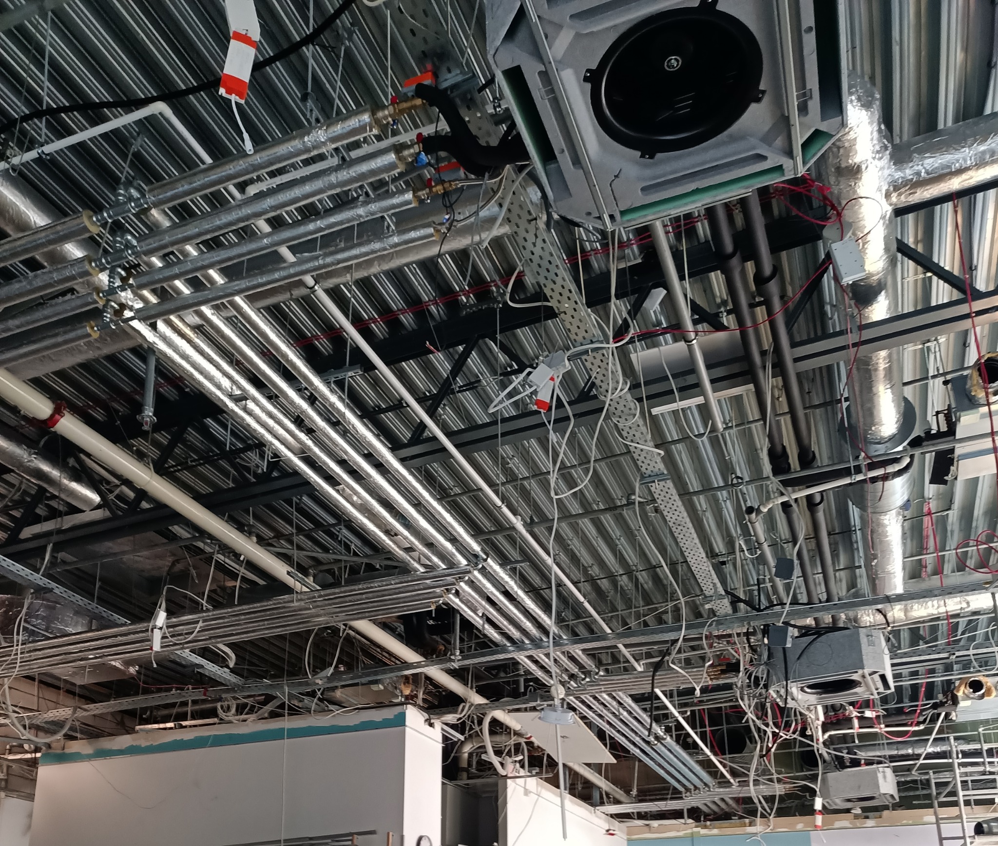 Widok na sufit w trakcie instalacji: liczne rury, kanały wentylacyjne, kable i elementy mocujące, z widocznymi fragmentami konstrukcji budynku.