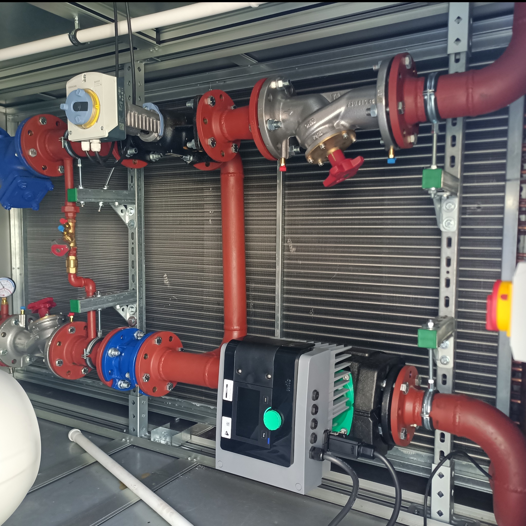 Instalacja grzewcza z czerwonymi rurami i niebieskimi elementami, widoczny wymiennik ciepła oraz pompa obiegowa z zielonym pokrętłem, całość zamontowana na metalowej konstrukcji.