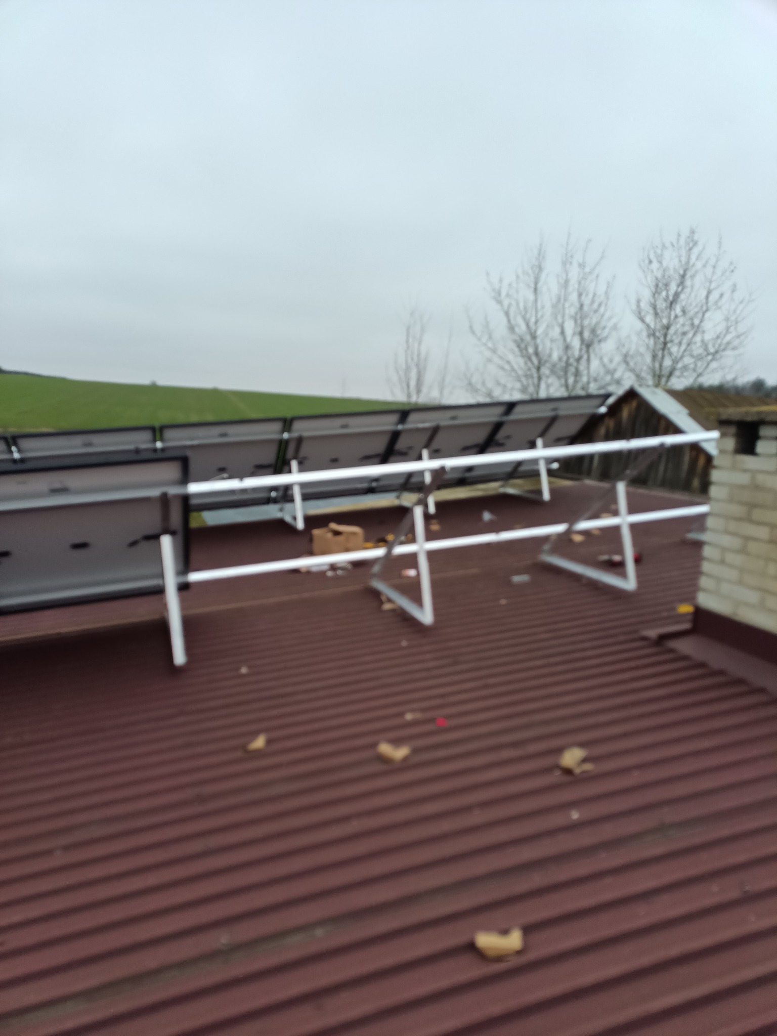 Montaż paneli fotowoltaicznych na brązowym, blaszanym dachu domu jednorodzinnego w pochmurny dzień, widoczne elementy konstrukcyjne i fragmenty kartonów po opakowaniach.