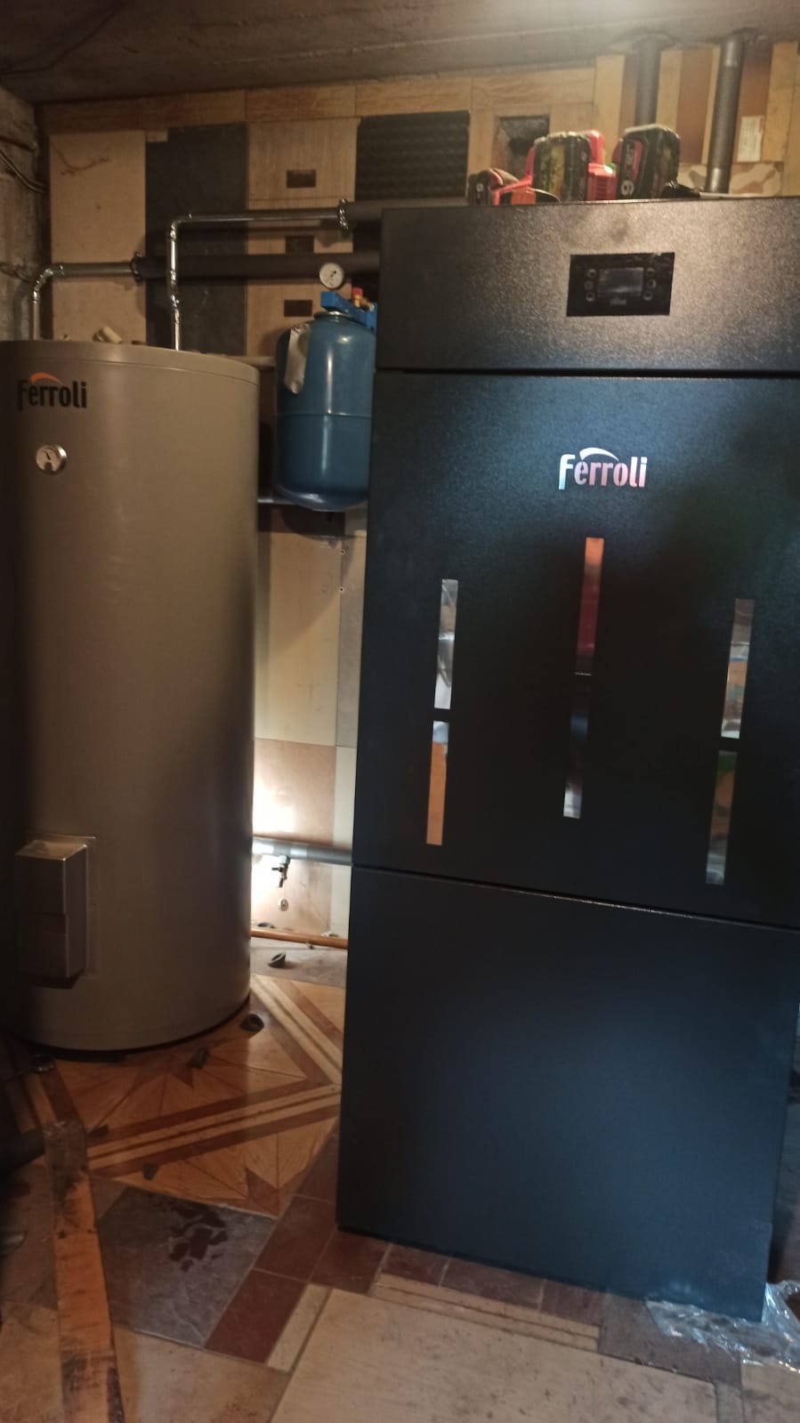 Zamontowany, czarny piec CO marki Ferroli z widocznym zbiornikiem buforowym oraz niebieskim naczyniem przeponowym w kotłowni z patchworkową ścianą i wzorzystą podłogą.