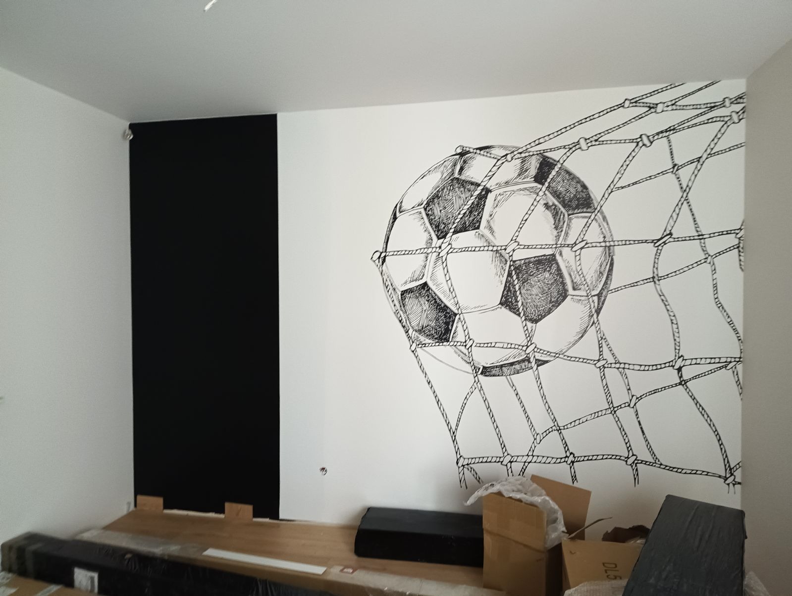 Ręcznie malowany mural piłki nożnej w siatce na białej ścianie w Rościszewie, obok pionowy czarny panel, kartony i materiały budowlane na podłodze.