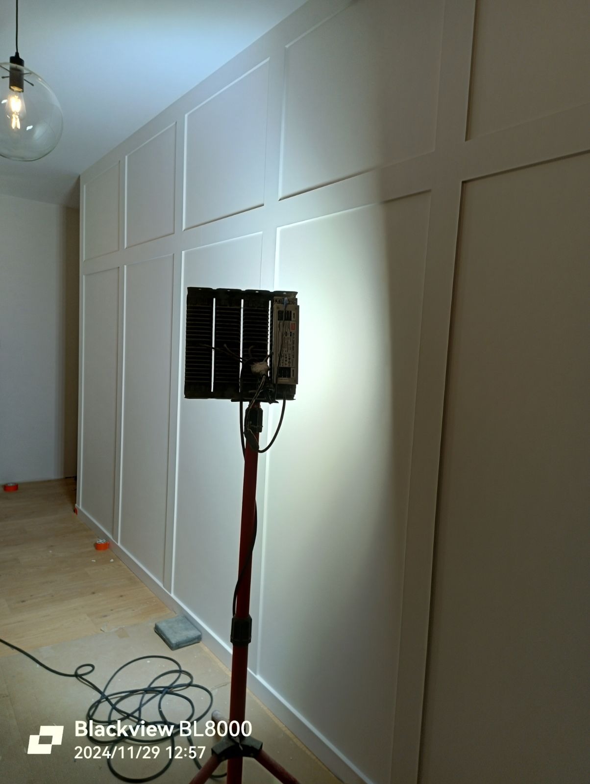 Świeżo pomalowana ściana z dekoracyjnymi panelami, oświetlona lampą roboczą na statywie. Widoczne kable i podkładki zabezpieczające podłogę.