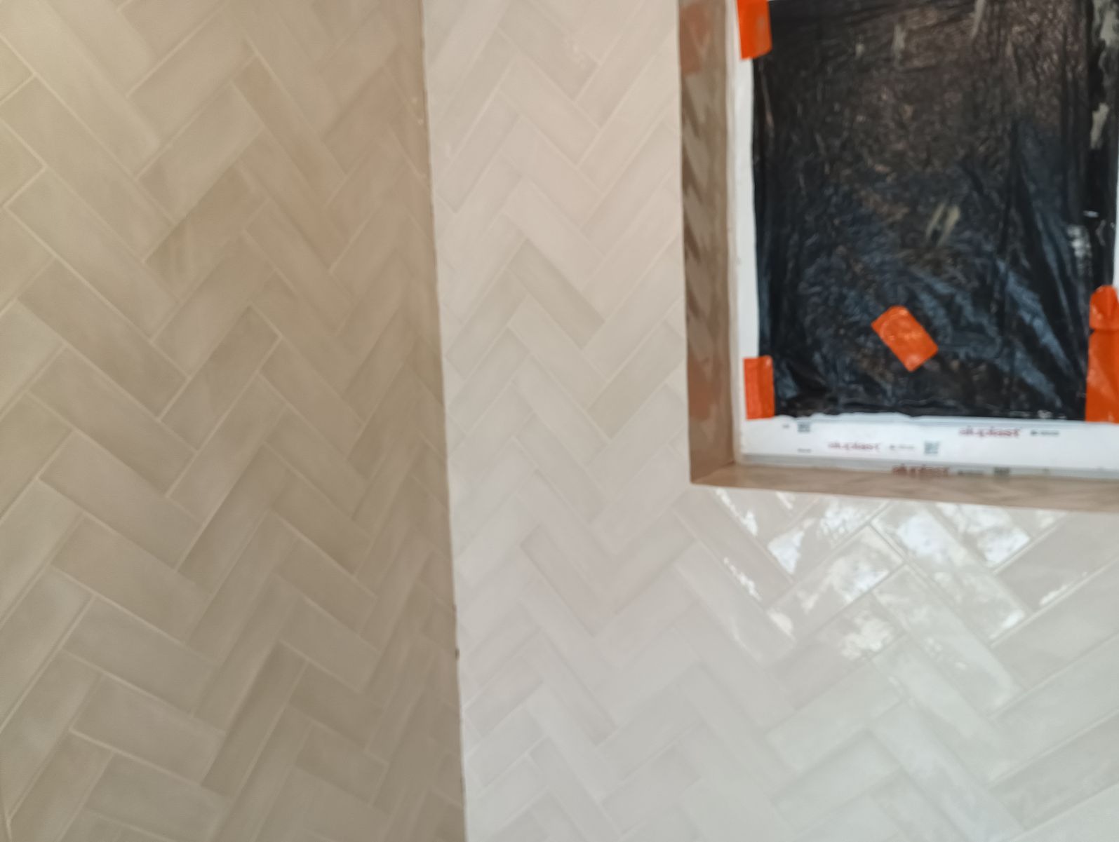 Ściana wyłożona błyszczącymi, kremowymi kafelkami w jodełkę, z widocznym oknem zabezpieczonym folią i taśmą malarską podczas prac wykończeniowych.