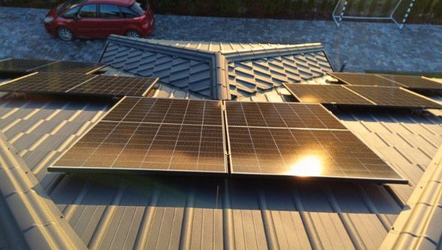 Panele fotowoltaiczne zamontowane na blaszanym dachu z widocznym odbiciem słońca, w tle czerwony samochód i fragment boiska.