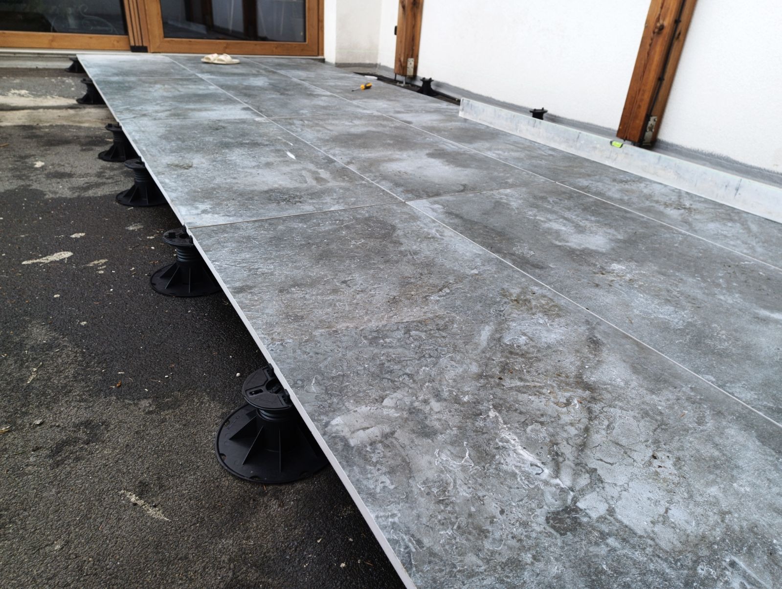 Montaż szarej, marmurkowej terakoty na regulowanych wspornikach tarasowych, widoczne narzędzia i poziomica, prace w toku.