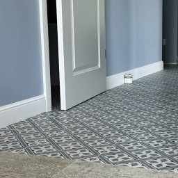 MAXIWIT SPÓŁKA Z OGRANICZONĄ ODPOWIEDZIALNOŚCIĄ - Ułożona, wzorzysta podłoga z płytek w odcieniach szarości i bieli, z widoczną listwą przypodłogową i fragmentem otwartych drzwi w pomieszczeniu o błękitnych ścianach.