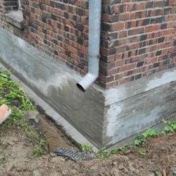 MAXIWIT SPÓŁKA Z OGRANICZONĄ ODPOWIEDZIALNOŚCIĄ - Odnowiona podmurówka budynku z czerwonej cegły, z widoczną rurą spustową i świeżo nałożoną warstwą szarego betonu, kontrastującą z ziemią i zielenią w tle.