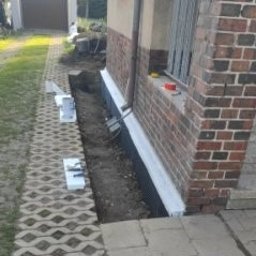 MAXIWIT SPÓŁKA Z OGRANICZONĄ ODPOWIEDZIALNOŚCIĄ - Odkopany fundament budynku z izolacją pionową, widoczne bloczki betonowe i kostka brukowa na zewnątrz. Prace ziemne w toku, przygotowanie do dalszych etapów izolacji.