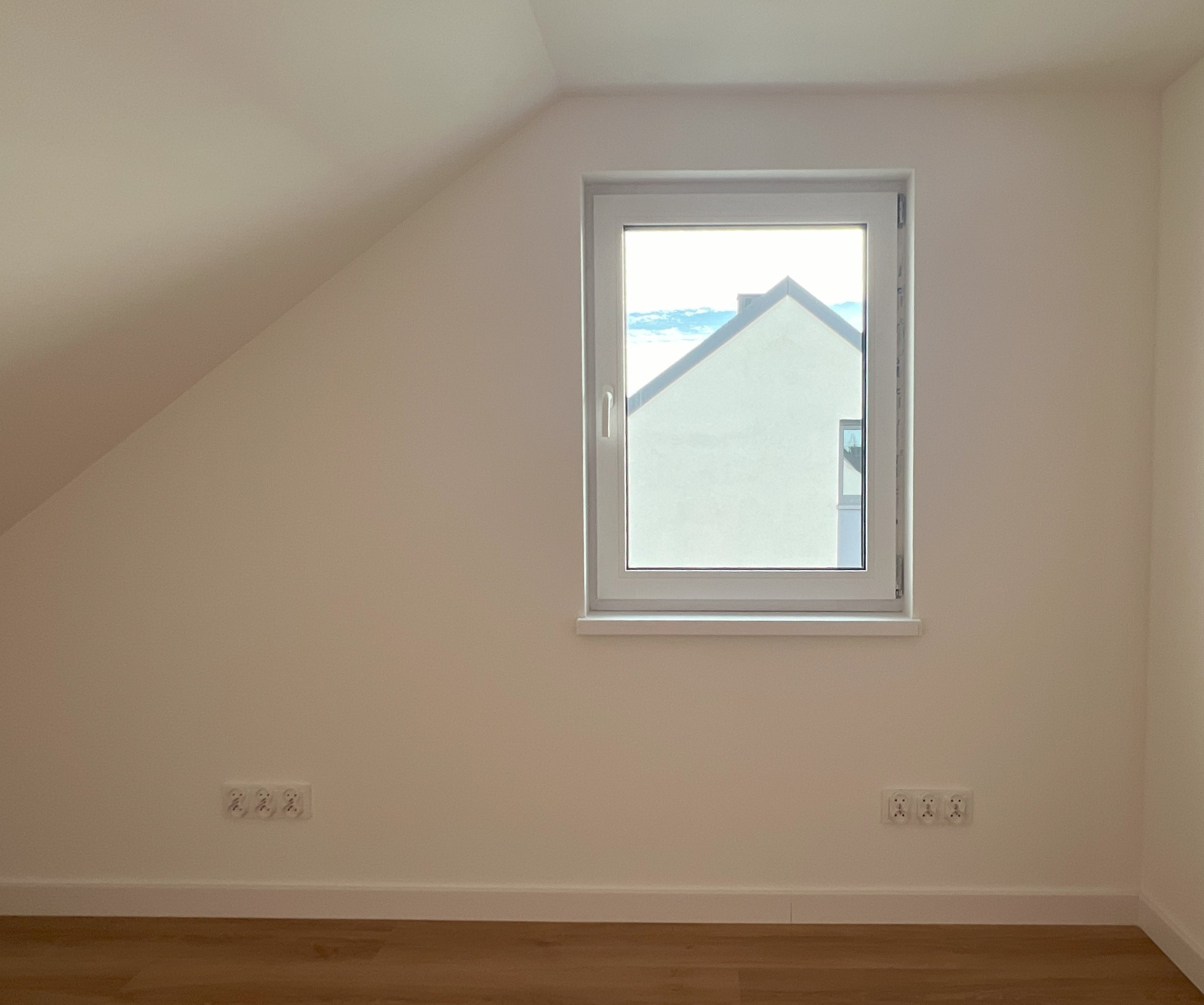 Wnętrze pokoju na poddaszu z nowo wstawionym oknem PCV, widok na biały budynek z niebieskim niebem w tle, drewnopodobne panele na podłodze, białe ściany z gniazdkami elektrycznymi.