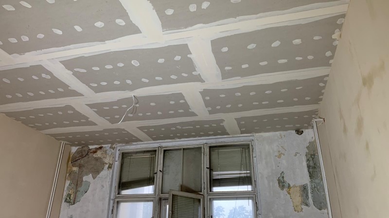 Sufit podwieszany z płyt kartonowo-gipsowych w trakcie montażu, widoczne łączenia płyt wzmocnione taśmą i szpachlą, ściany w różnym stanie wykończenia, okno z żaluzjami.