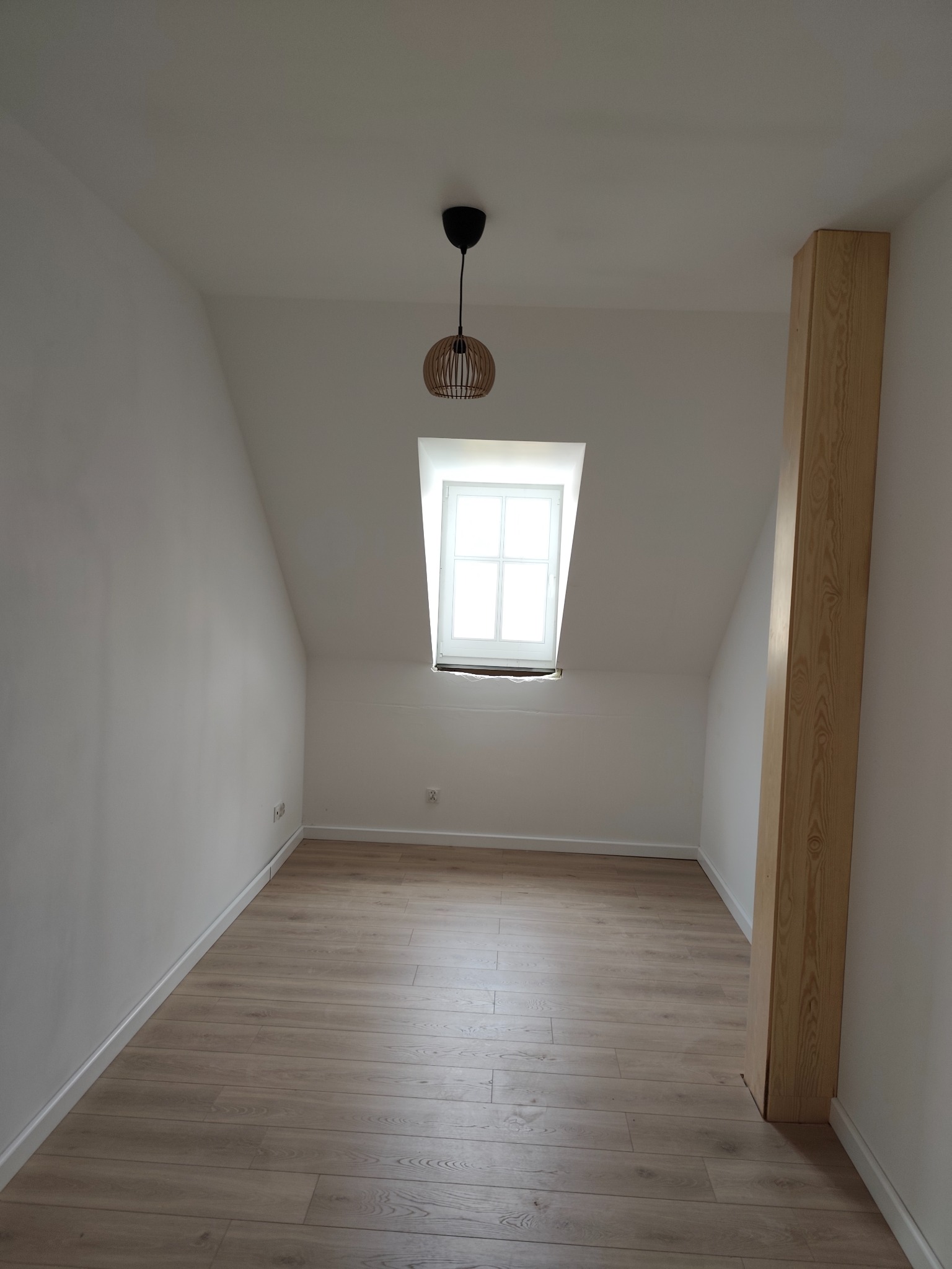 Jasne, wykończone poddasze z drewnianą podłogą, białymi ścianami i oknem w szczycie. Widoczna lampa wisząca i drewniany element konstrukcyjny. Minimalistyczny styl.