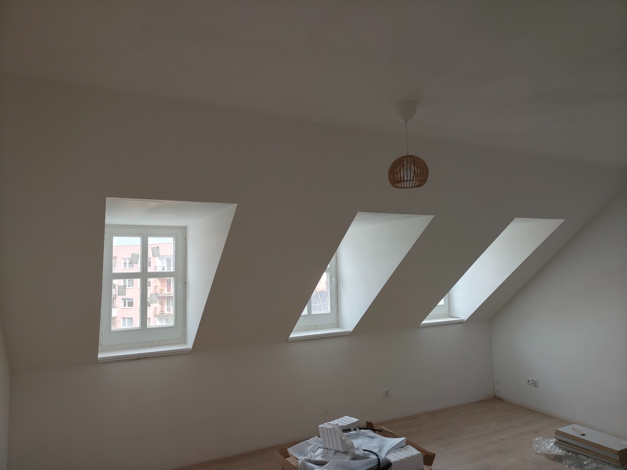 Jasne poddasze po remoncie z trzema oknami dachowymi, drewnianą podłogą i lampą wiszącą. Na podłodze kartony z materiałami budowlanymi.