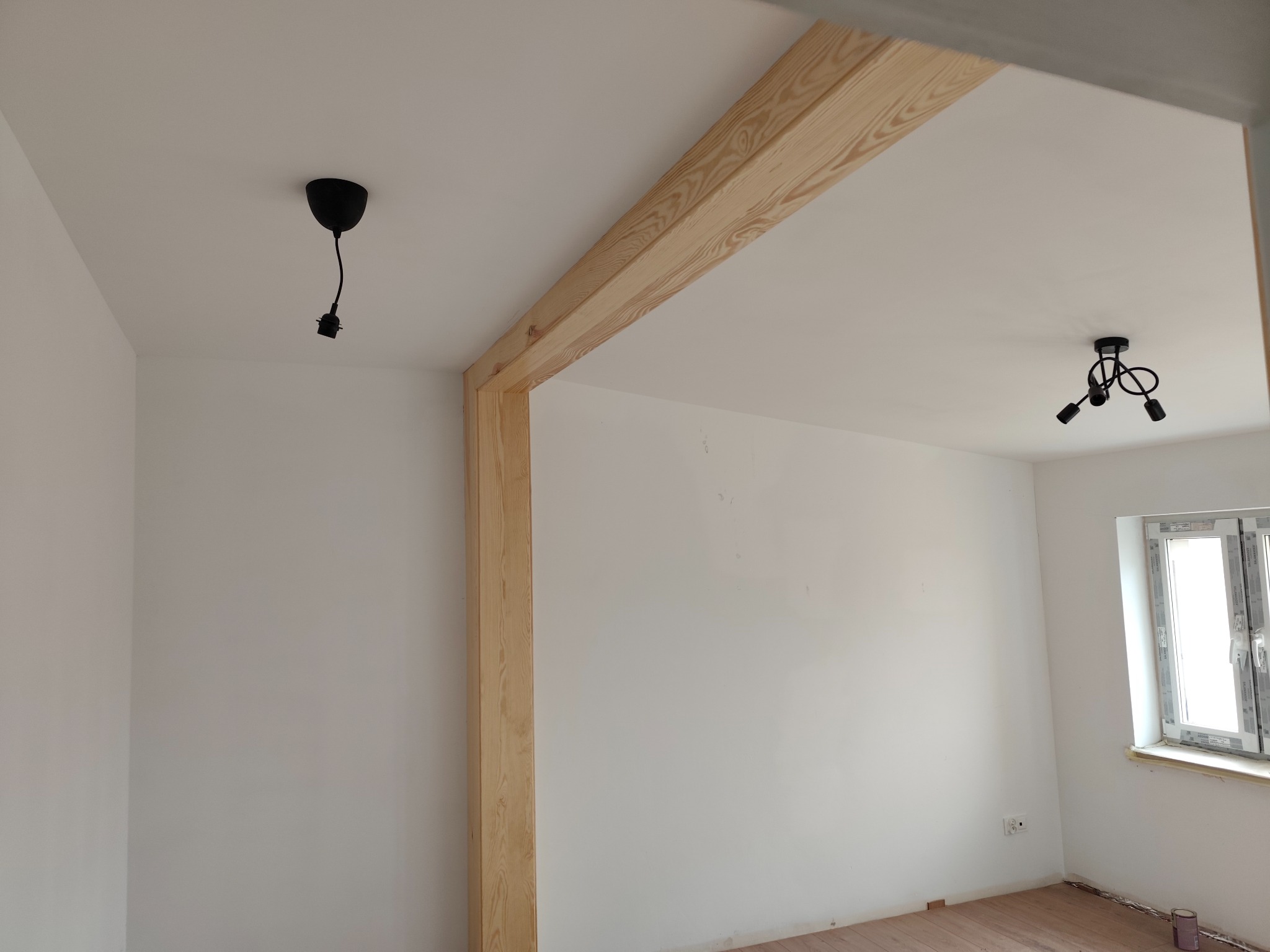 Jasne drewniane obramowanie drzwi w białym pokoju z parkietem. Widoczne czarne lampy sufitowe i okno. Minimalistyczny design wnętrza po remoncie.