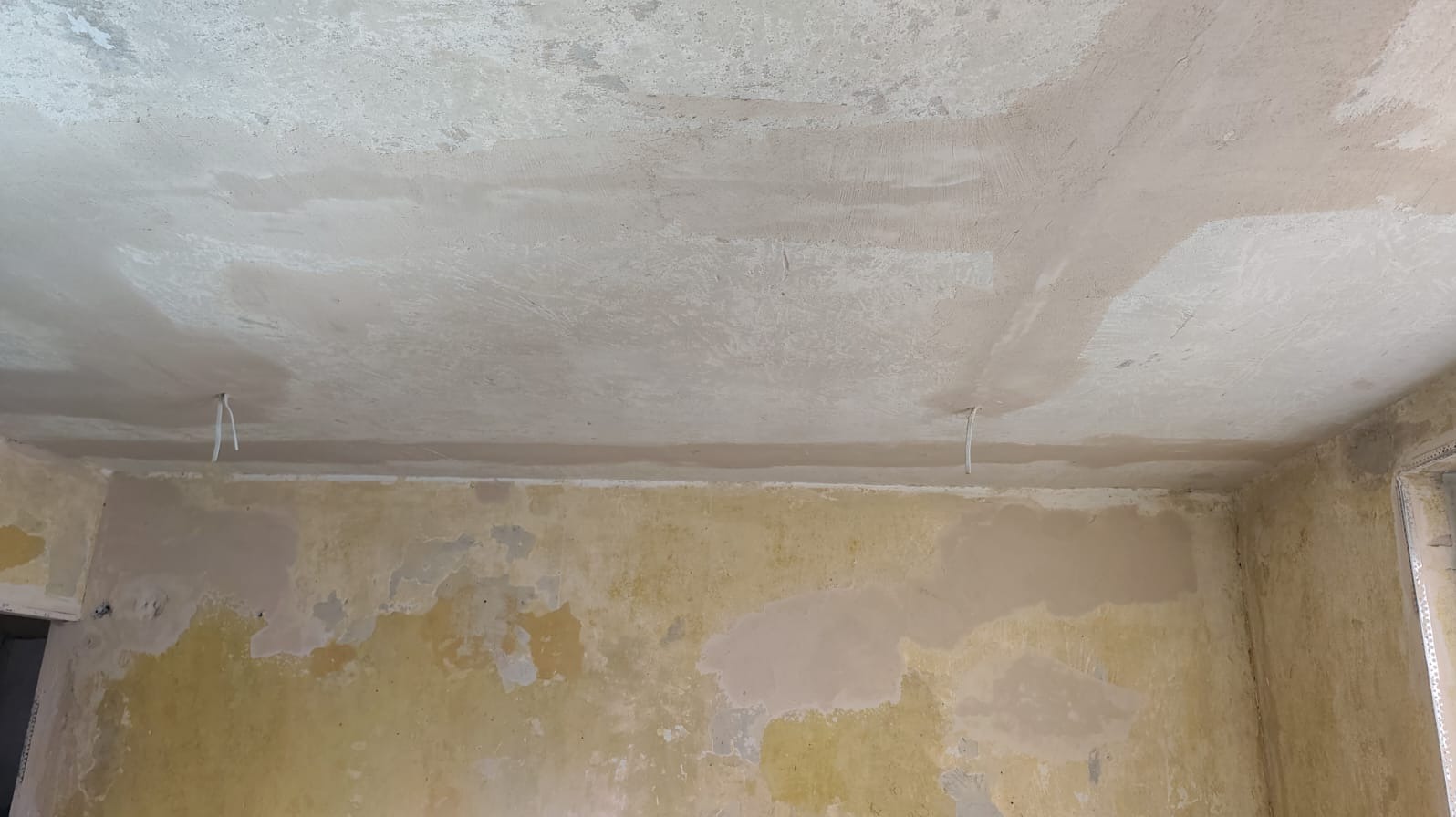 Przygotowanie ścian i sufitu do szpachlowania: częściowo zagruntowane, z widocznymi śladami starej farby i wystającymi przewodami elektrycznymi. Widok na róg pomieszczenia.