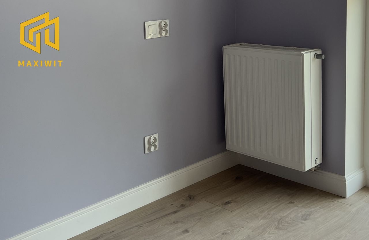 Szary pokój po remoncie: świeżo pomalowana ściana z dwoma gniazdkami, biały grzejnik i listwa przypodłogowa, panele podłogowe. Logo firmy w lewym górnym rogu.