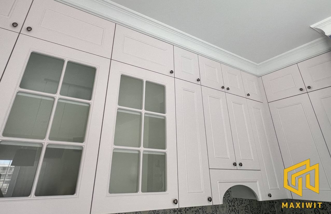 Jasne, klasyczne szafki kuchenne z przeszklonymi frontami i ozdobnymi listwami, widoczne od dołu pod kątem. Ciemne okucia i wzorzysta ściana nad blatem.