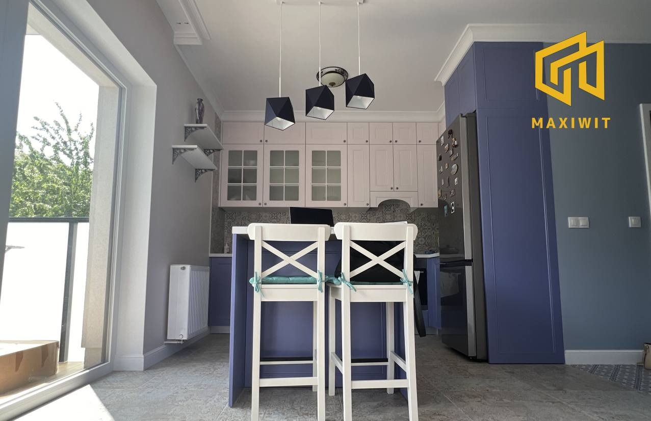 Nowoczesna kuchnia w odcieniach bieli i fioletu z wyspą i hokerami. Widoczne wiszące lampy i dekoracyjne półki. Ujęcie z perspektywy wejścia, z oknem po lewej stronie.