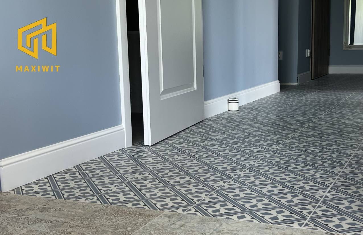 Ułożona, wzorzysta podłoga z płytek w odcieniach szarości i bieli, z widoczną listwą przypodłogową i fragmentem otwartych drzwi w pomieszczeniu o błękitnych ścianach.