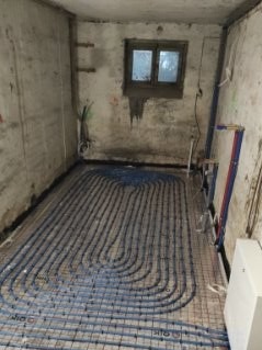 Instalacja ogrzewania podłogowego w surowym pomieszczeniu z oknem. Rury ułożone w pętle na siatce, widoczne ślady po remoncie na ścianach.