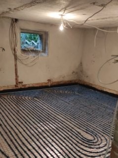 Ułożone rury ogrzewania podłogowego w surowym pomieszczeniu z oknem. Widoczne kable na ścianie i surowy beton. Etap prac przed wylaniem posadzki.