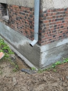 Odnowiona podmurówka budynku z czerwonej cegły, z widoczną rurą spustową i świeżo nałożoną warstwą szarego betonu, kontrastującą z ziemią i zielenią w tle.